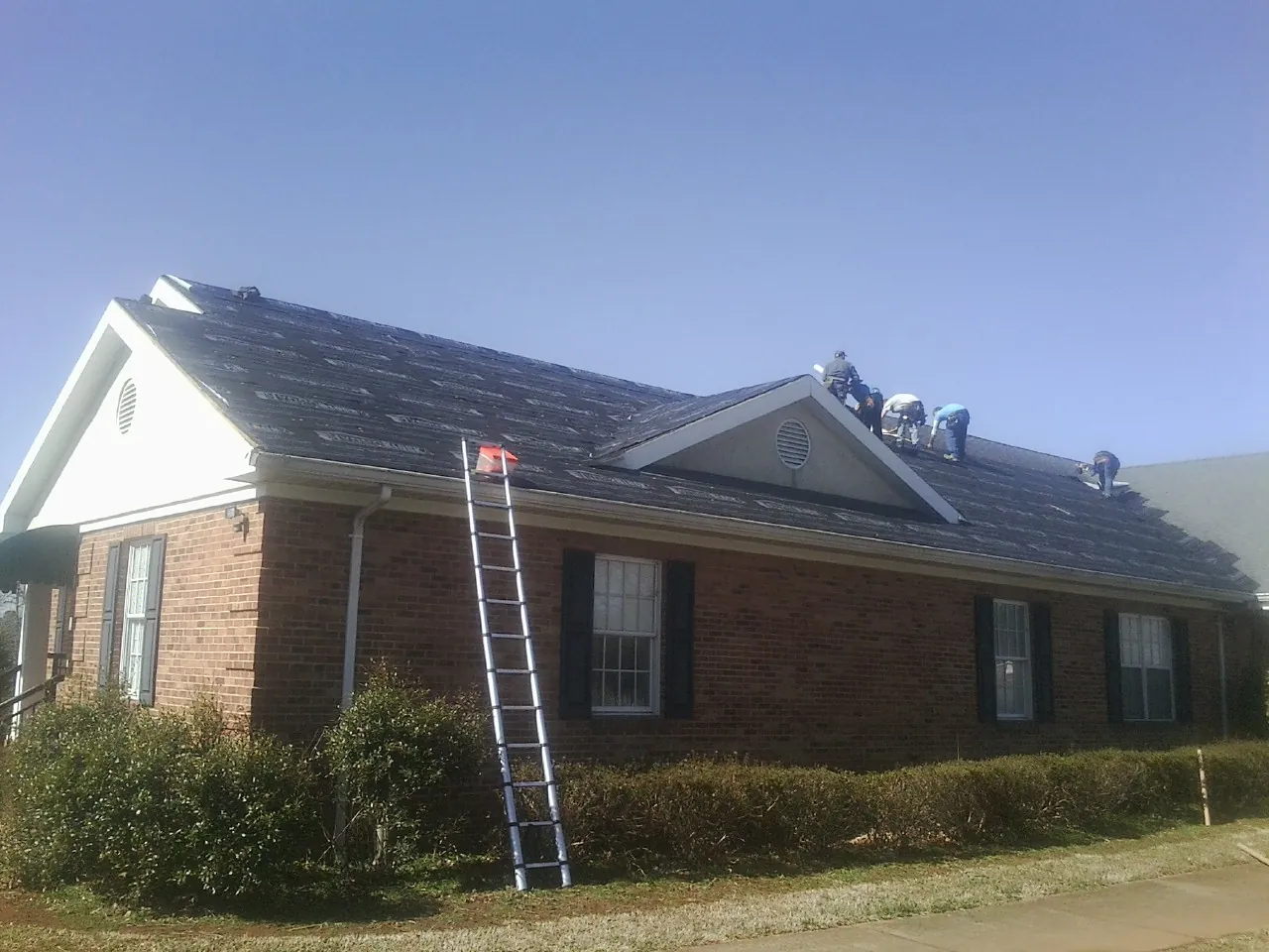 Crew tearing shingles off a brick ranch roof with a ladder set — Metal Roof Repair work by our crew in Weldona
