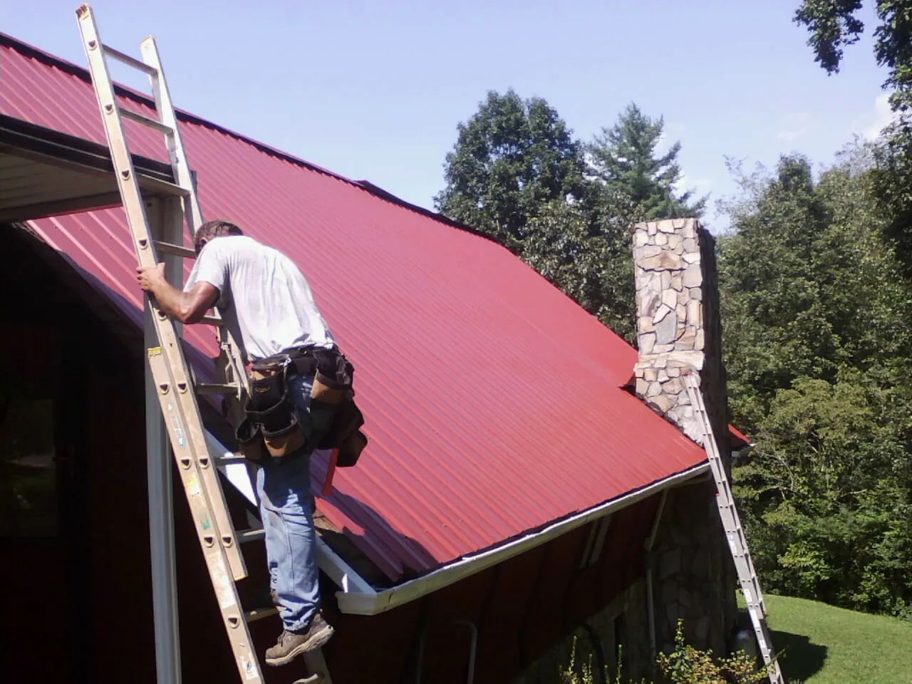 Roofer on a ladder beside a red metal roof and stone chimney — Soffit & Fascia Repair by Roofing Brothers in Weldona